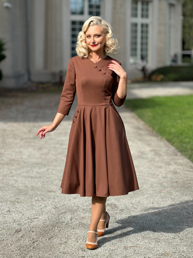 Modelka w cynamonowej sukience Vivian z kopertowym dekoltem, dopasowaną talią i rozkloszowanym dołem, stojąca przed jasnym budynkiem.