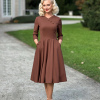 Modelka w cynamonowej sukience Vivian z kopertowym dekoltem i rozkloszowanym dołem, trzymająca dłonie w bocznych kieszeniach.