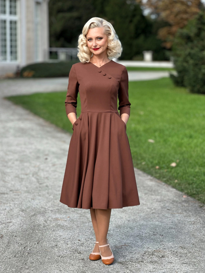 Modelka w cynamonowej sukience Vivian z kopertowym dekoltem i rozkloszowanym dołem, trzymająca dłonie w bocznych kieszeniach.